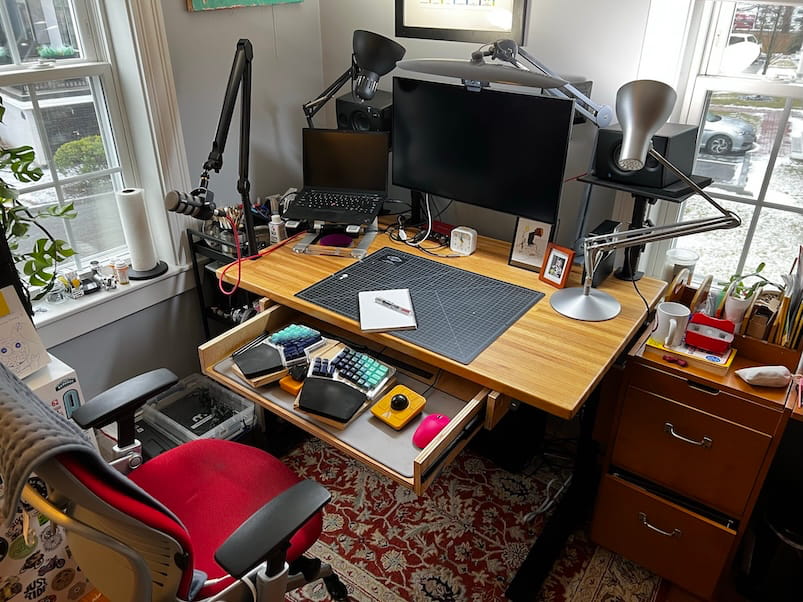 A busy desk with drawers pulled open, showing keyboards and accessories
inside. The desktop holds a cutting mat, notebook, monitor, and desk lamps, with
plants and storage units nearby.
