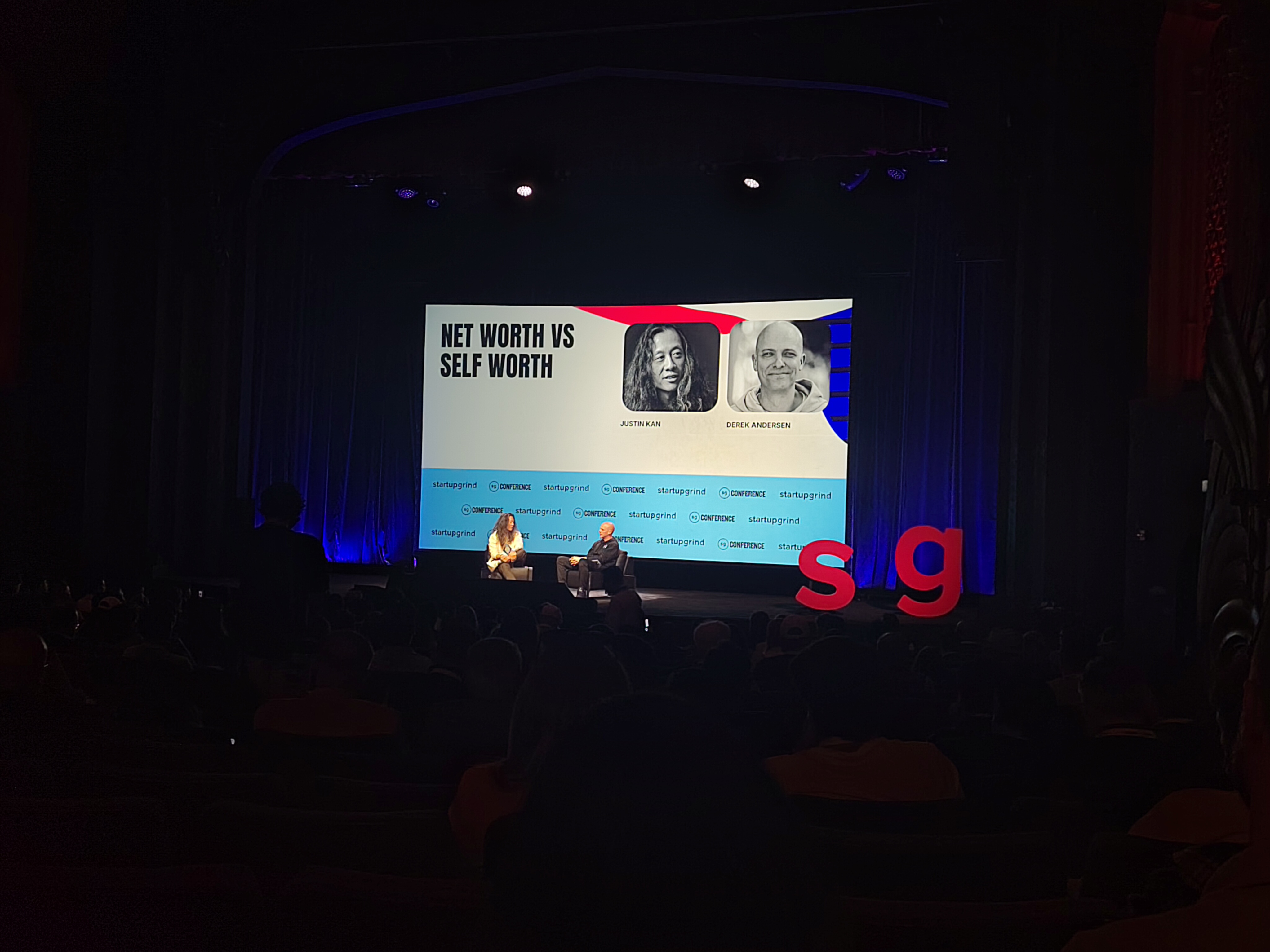 A dark auditorium at the startup grind conference. Two men are on stage with a large screen behind them and a small "s" and "g" red sign on the right of the stage
