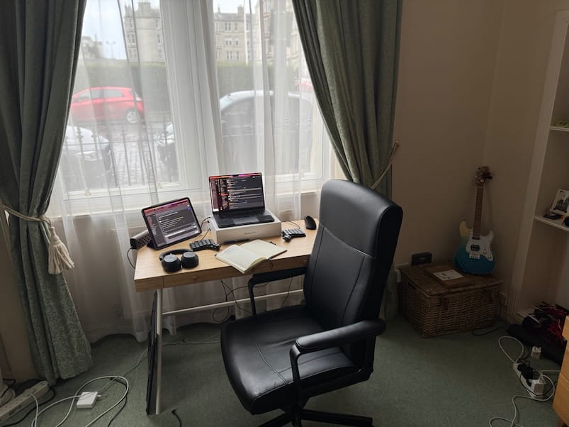 A compact home office setup by a large window, with a small desk holding a
laptop, external keyboard, notebook, and headphones. A black office chair faces
the desk, and daylight fills the room.