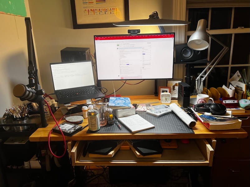 A cluttered creative workspace with a wooden desk covered in notebooks, tools,
cables, mugs, and stationery. A large monitor and laptop sit at the back, with
desk lamps and shelves surrounding the area.