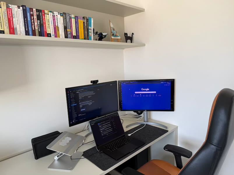 A clean white desk setup with two external monitors and a laptop on a stand. A
keyboard, mouse, and desk mat sit below, with books and small decorative objects
on a shelf above.