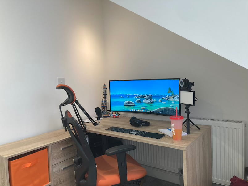 A modern home office desk under a sloped ceiling, featuring a wide monitor,
keyboard, headphones, microphone arm, and a pink drinks bottle. An ergonomic
chair sits in front of the desk.