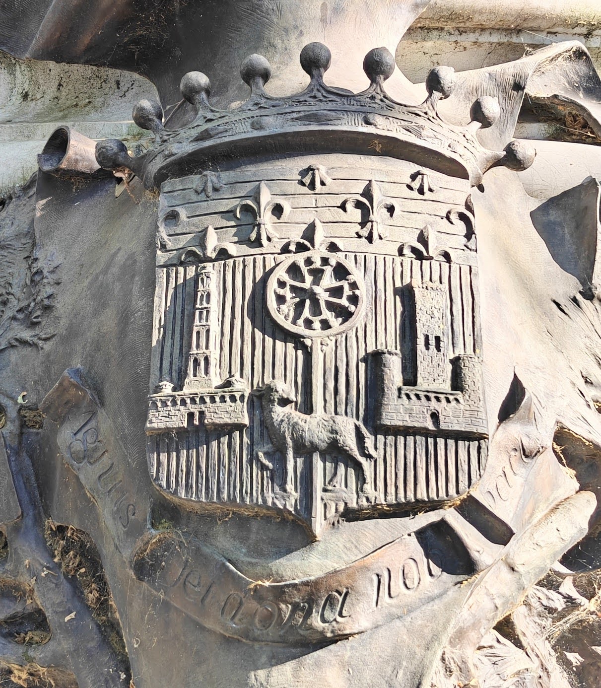Bronze relief of a coat of arms in harsh side light: a shield with vertical bars, a row of fleurs-de-lys across the top, a lamb, a cross inside a circle, and two towers, above a scroll bearing a worn Latin motto
